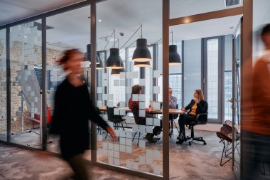 İş ortamının hareketli koşuşturmasında, bir grup genç iş adamı ofislerinin yanındaki koridorda yürürken, meslektaşları içeride iş birliği yaparak,