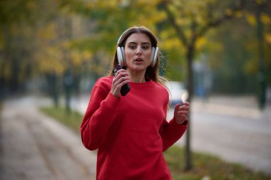Sağlıklı yaşam tarzı, sabah koşusu. Genç ve güzel bir kadın sonbahar parkında koşuyor ve akıllı telefonuyla kulaklıkla müzik dinliyor. Kırmızı bir spor gömlek giyiyor.. 