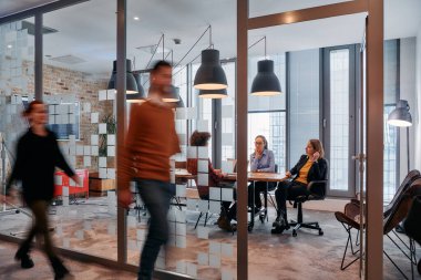 İş ortamının hareketli koşuşturmasında, bir grup genç iş adamı ofislerinin yanındaki koridorda yürürken, meslektaşları içeride iş birliği yaparak,