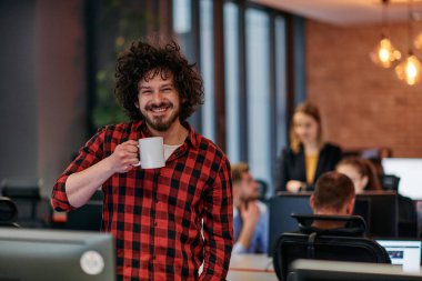 Bilgisayarlarla çevrili bir ofis ortamında ilgi çekici bir işadamı, elinde kahve fincanı tutarken görevlerine odaklanıyor, konsantrasyon ve verimliliği özetliyor.. 