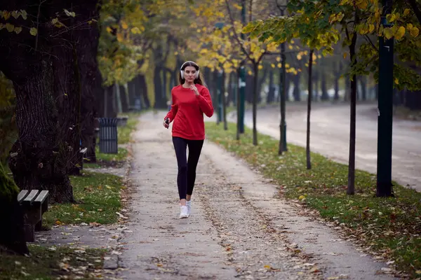 Sağlıklı yaşam tarzı, sabah koşusu. Genç ve güzel bir kadın sonbahar parkında koşuyor ve akıllı telefonuyla kulaklıkla müzik dinliyor. Kırmızı bir spor gömlek giyiyor.. 