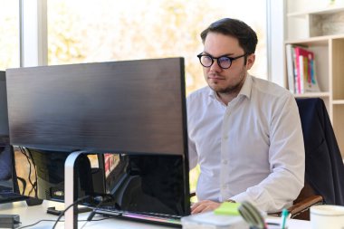 Modern saç stiline sahip beyaz gömlekli bir şirket yöneticisi, şık ve profesyonel bir ofis ortamında bilgisayarına odaklanıyor..