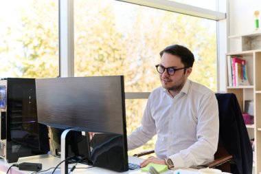 Modern saç stiline sahip beyaz gömlekli bir şirket yöneticisi, şık ve profesyonel bir ofis ortamında bilgisayarına odaklanıyor..