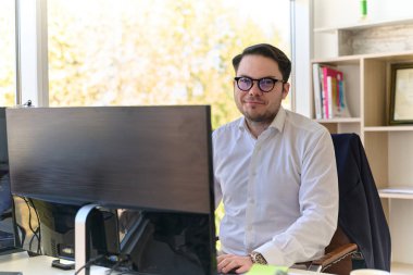 Modern saç stiline sahip beyaz gömlekli bir şirket yöneticisi, şık ve profesyonel bir ofis ortamında bilgisayarına odaklanıyor..