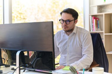 Modern saç stiline sahip beyaz gömlekli bir şirket yöneticisi, şık ve profesyonel bir ofis ortamında bilgisayarına odaklanıyor..