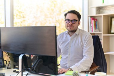 Modern saç stiline sahip beyaz gömlekli bir şirket yöneticisi, şık ve profesyonel bir ofis ortamında bilgisayarına odaklanıyor..