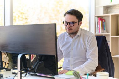 Modern saç stiline sahip beyaz gömlekli bir şirket yöneticisi, şık ve profesyonel bir ofis ortamında bilgisayarına odaklanıyor..
