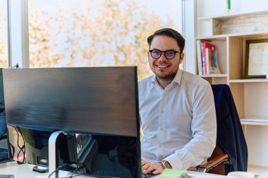 Modern saç stiline sahip beyaz gömlekli bir şirket yöneticisi, şık ve profesyonel bir ofis ortamında bilgisayarına odaklanıyor..