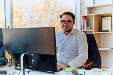 Modern saç stiline sahip beyaz gömlekli bir şirket yöneticisi, şık ve profesyonel bir ofis ortamında bilgisayarına odaklanıyor..