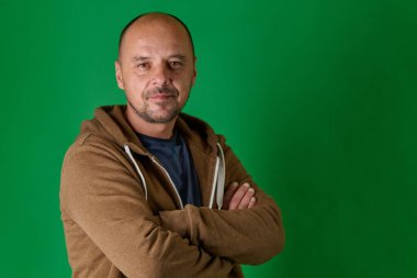 Sıradan giyinmiş orta yaşlı kel bir adam kollarını kavuşturmuş ve rahat bir gülümseme ile canlı yeşil bir stüdyo arka planına kurulmuş.