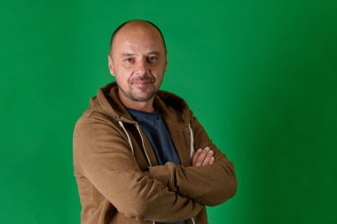 Sıradan giyinmiş orta yaşlı kel bir adam kollarını kavuşturmuş ve rahat bir gülümseme ile canlı yeşil bir stüdyo arka planına kurulmuş.
