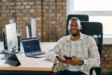  Afrikalı Amerikalı girişimci, sosyal medyayı taramak için akıllı telefon kullanarak modern bir ofise ara veriyor, iş çabaları arasında dijital bir bağlantı ve rahatlama anı yakalıyor