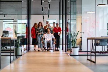 A diverse group of young business people walking a corridor in the glass-enclosed office of a modern startup, including a person in a wheelchair and a woman wearing a hijab. clipart