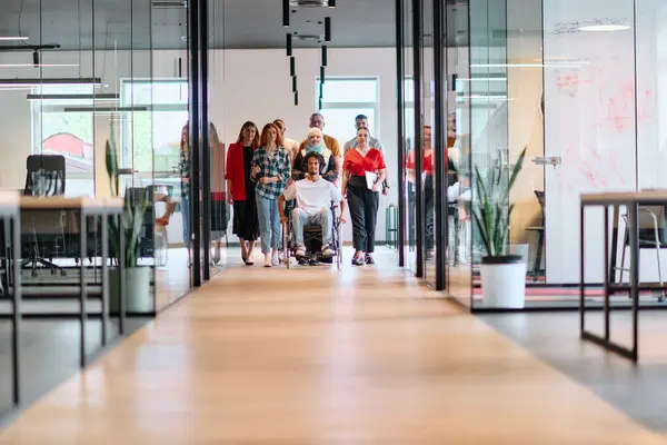 stock image A diverse group of young business people walking a corridor in the glass-enclosed office of a modern startup, including a person in a wheelchair and a woman wearing a hijab.