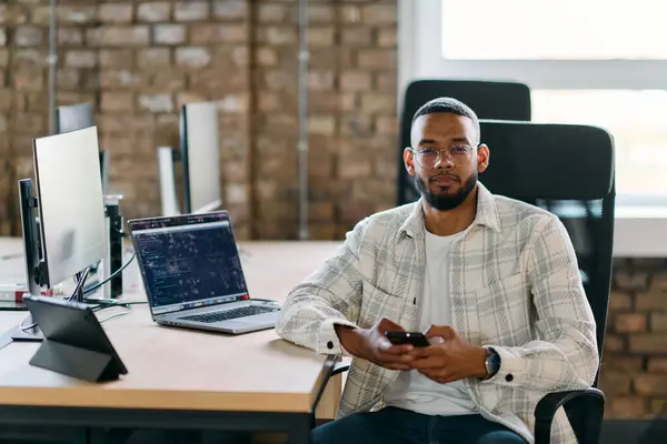 stock image  African American entrepreneur takes a break in a modern office, using a smartphone to browse social media, capturing a moment of digital connectivity and relaxation amidst his business endeavors