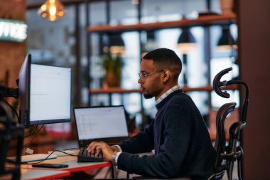 Yetenekli bir Afrikalı Amerikalı programcı, modern bir ofiste bilgisayarının başına oturmuş, problem çözme ve teknoloji konusundaki uzmanlığını sergiliyor..