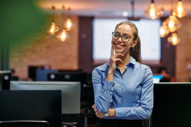 Mavi gömlekli göz kamaştırıcı bir iş kadını ofisinin önünde duruyor, parlak ve çekici bir gülümsemeyle kararlılığını gösteriyor..