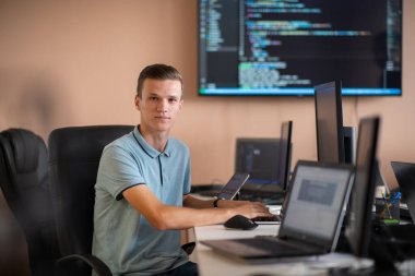 Genç bir programcı ofisinde masasında oturmuş, akıllı telefonlar için yenilikçi yeni uygulamaları test etmeye odaklanmış, yaratıcılık ve teknoloji ruhunu somutlaştırıyor..