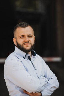 Tişörtlü başarılı genç bir iş adamı, kollarını kavuşturmuş, açık havada poz veriyor, yüzünde kendinden emin bir ifade var.