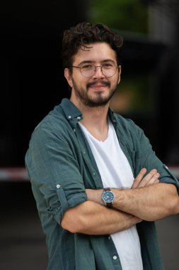 Tişörtlü başarılı genç bir iş adamı, kollarını kavuşturmuş, açık havada poz veriyor, yüzünde kendinden emin bir ifade var.