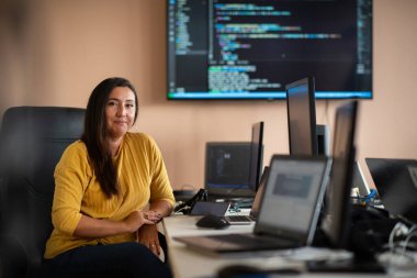Bilgisayarlarla çevrili bir programcı ofisinde oturan bir iş kadını uzmanlığını ve teknolojiye olan bağlılığını gösteriyor.