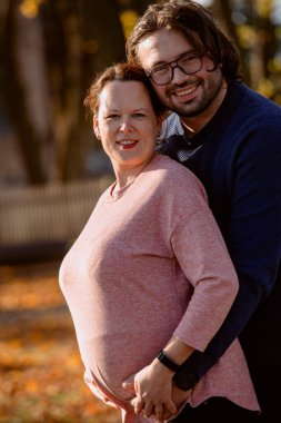 Bir adam parkta hamile karısını sevgiyle kucaklıyor. Huzurlu bir sıcaklık ve şefkat anı yaşıyor..