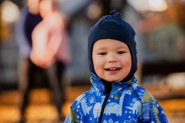 A joyful little boy in his winter coat and hat plays happily in the park, surrounded by the love and warmth of his smiling parents. clipart