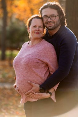 Bir adam parkta hamile karısını sevgiyle kucaklıyor. Huzurlu bir sıcaklık ve şefkat anı yaşıyor..