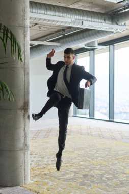 Modern bir ofise kendinden geçmiş bir şekilde atlayan bir işadamı, elinde bir evrak çantasıyla, başarı ve zafer anını kutluyor.