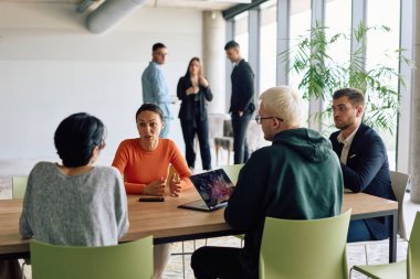 A diverse team of business professionals engaged in a discussion around a conference table in an office, while their colleagues collaborate in the background.  clipart