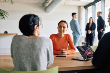 A diverse team of business professionals engaged in a discussion around a conference table in an office, while their colleagues collaborate in the background.  clipart