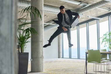 Modern bir ofise kendinden geçmiş bir şekilde atlayan bir işadamı, elinde bir evrak çantasıyla, başarı ve zafer anını kutluyor.