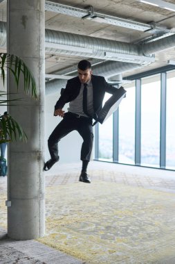 Modern bir ofise kendinden geçmiş bir şekilde atlayan bir işadamı, elinde bir evrak çantasıyla, başarı ve zafer anını kutluyor.