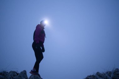 Kadın yürüyüşçü ya da triatlon patika koşucusu. Gece sisle dağda bir patikada farlı..