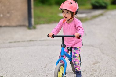 Şirin bir kostüm giymiş ve kask takan küçük bir kız heyecanlı bir şekilde ilk kez bisiklet sürmeyi öğreniyor, neşe ve kararlılıkla dolu.
