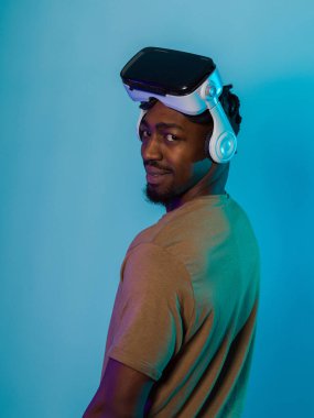 In a futuristic visual, an African American man stands isolated against a striking blue backdrop, adorned with VR glasses that transport him into a cutting-edge virtual reality experience, merging clipart