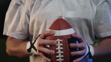 Takım üniformalı, kasklı, futbol topu tutan, başını kaldıran ve sinematik yavaş çekimde kameraya güvenle bakan Amerikalı bir futbolcunun yakın çekimi.