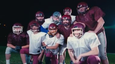 Bir grup Amerikan futbolcusu, kestane rengi ve beyaz üniformalı, kasklı ve koruyucu kıyafetli bir grup, gece bir sahada birlikte poz veriyorlar. Takım, stadyumun altında dostluk ve takım çalışması gösteriyor.