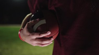 Stadyum ışıkları altında topu tutan, bordo forma giyen bir Amerikan futbolcusunun yakın çekim görüntüsü. Spor terfileri ya da tasarımlar için mükemmel.