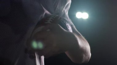 Takım üniformalı, kasklı, futbol topu tutan, başını kaldıran ve sinematik yavaş çekimde kameraya güvenle bakan Amerikalı bir futbolcunun yakın çekimi.
