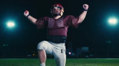 Amerikalı futbolcu, dramatik ışıklar altında zaferi kutluyor ve kollarını zaferle kaldırıyor. Zor bir maçın ardından güçlü bir zafer, heyecan ve takım ruhu anı..