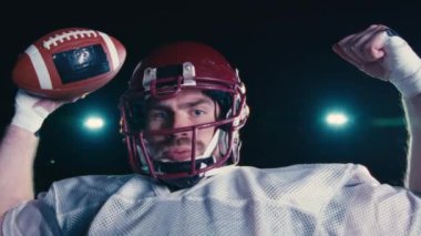 Amerikalı futbolcu, dramatik ışıklar altında zaferi kutluyor ve kollarını zaferle kaldırıyor. Zor bir maçın ardından güçlü bir zafer, heyecan ve takım ruhu anı..