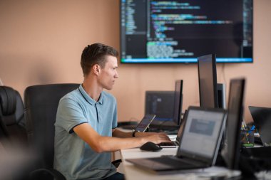 Genç bir programcı ofisinde masasında oturmuş, akıllı telefonlar için yenilikçi yeni uygulamaları test etmeye odaklanmış, yaratıcılık ve teknoloji ruhunu somutlaştırıyor..