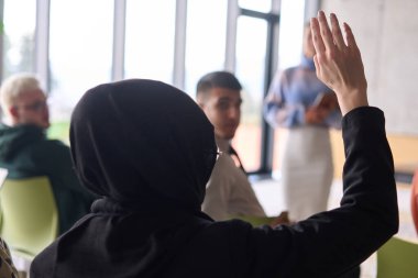 A Muslim businesswoman, dressed in professional attire and wearing a hijab, raises her hand during a business meeting to ask a question to the educator leading the session.  clipart