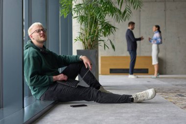 Modern bir ofiste geniş bir pencerenin yanında oturan sıradan bir profesyonel bilgisayarla çalışırken gülümseyen meslektaşları bulanık arka planda etkileşime giren biri.