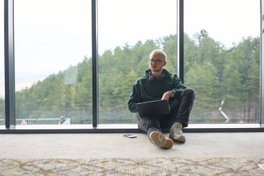 Modern bir ofisin zemininde oturan sarışın bir adam, kucağında dizüstü bilgisayarla pencereye yaslanmış, rahat ve odaklanmış bir tavır sergilerken tamamen işine odaklanmış.