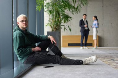 Modern bir ofiste geniş bir pencerenin yanında oturan sıradan bir profesyonel bilgisayarla çalışırken gülümseyen meslektaşları bulanık arka planda etkileşime giren biri.