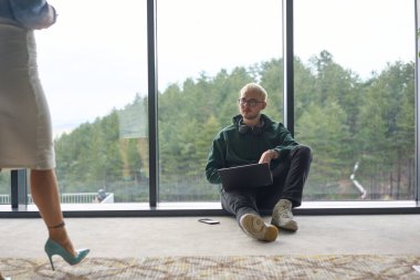 Sarışın bir adam modern bir ofisin zemininde rastgele oturuyor, dizüstü bilgisayarını kullanırken pencereye yaslanıyor, yanında çekici bayan arkadaşı duruyor..