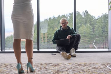 Sarışın bir adam modern bir ofisin zemininde rastgele oturuyor, dizüstü bilgisayarını kullanırken pencereye yaslanıyor, yanında çekici bayan arkadaşı duruyor..