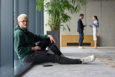 Modern bir ofiste geniş bir pencerenin yanında oturan sıradan bir profesyonel bilgisayarla çalışırken gülümseyen meslektaşları bulanık arka planda etkileşime giren biri.